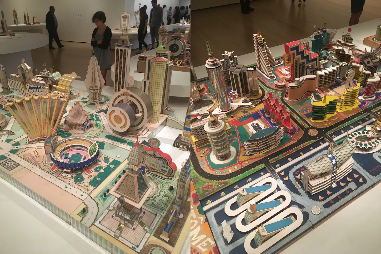 The image features two different views of intricate architectural models displayed in an exhibition space. The models are colorful and detailed, representing futuristic or imaginative cityscapes with a variety of building designs, including towering skyscrapers, rounded structures, and geometric patterns.