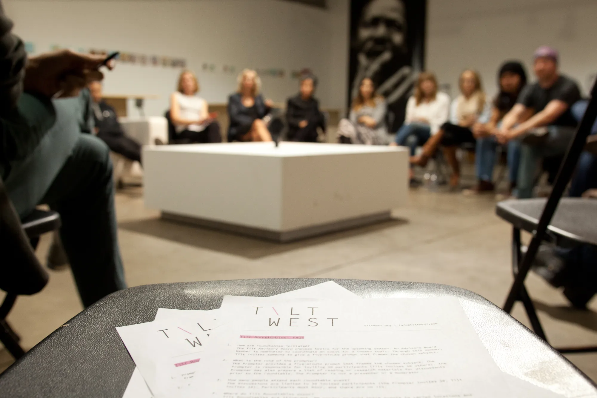 A group of people sit in a circle, slightly out of focus in the background, as they participate in a discussion. In the foreground, in focus, are papers on a chair with the title "TILT WEST" visible at the top of the documents.