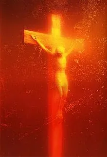 A photograph of a crucifix submerged in a bright red-orange liquid, possibly blood or a similar substance, giving the image an intense, glowing effect. The figure on the crucifix is illuminated, creating a dramatic and somewhat unsettling visual impact. 