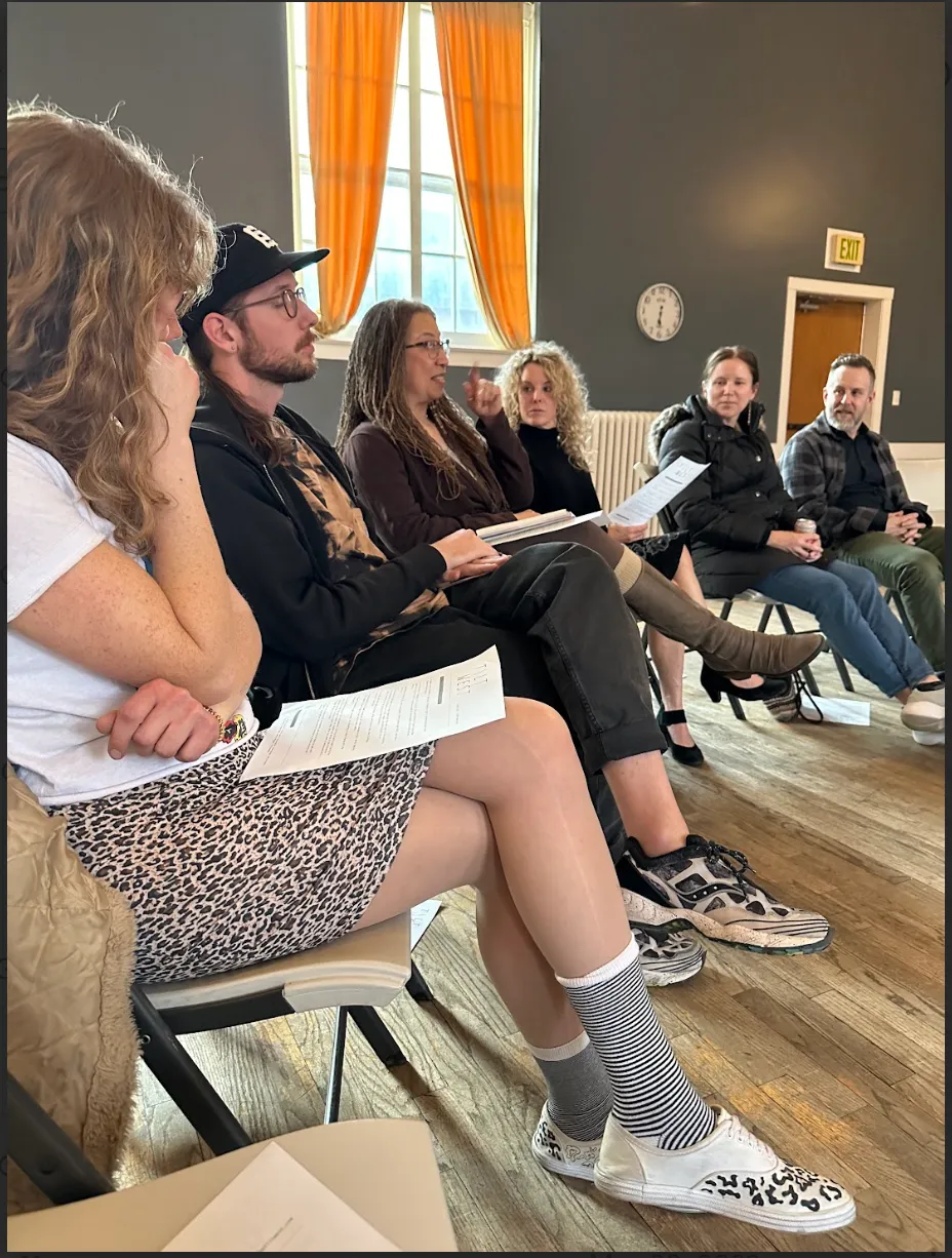 A group of people sit in a semi-circle in a room with wooden flooring and large windows covered with bright orange curtains. The person in the center, with long hair and glasses, is speaking while holding a paper, and the others listen attentively.