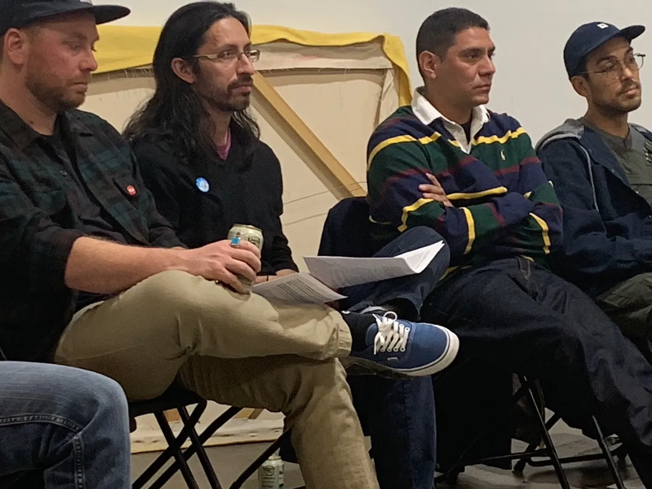 A group of four men sit in a row, attentively listening during a discussion. The background includes a partially visible canvas frame with a yellow covering.