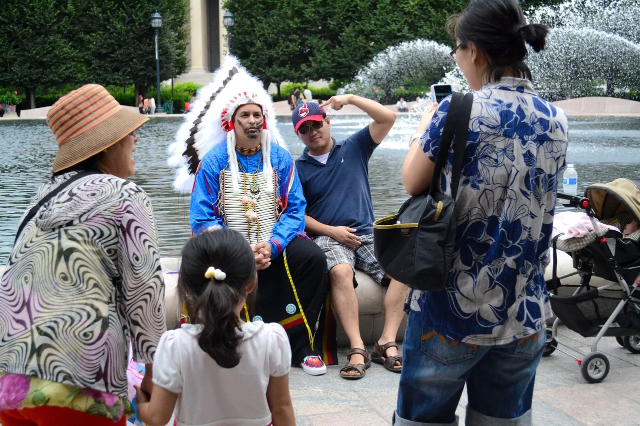 A man dressed in a blue shirt and an American Indian headdress sits by a fountain, posing for a photograph with a group of people. A man next to him, wearing a baseball cap, points at the headdress while smiling. A woman and child, both facing away from the camera, are also in the foreground. The fountain and trees in the background create a park-like setting.