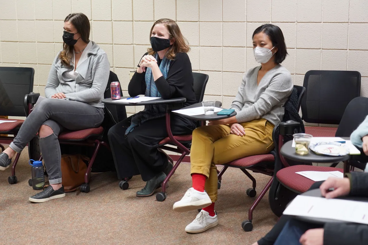 Three women sit in a row in a classroom setting, engaged in a discussion. All are wearing face masks. The woman on the left wears a gray hoodie and jeans, while the woman in the middle has a blue scarf and a black outfit. The woman on the right wears a gray sweater, mustard-yellow pants, and red socks.