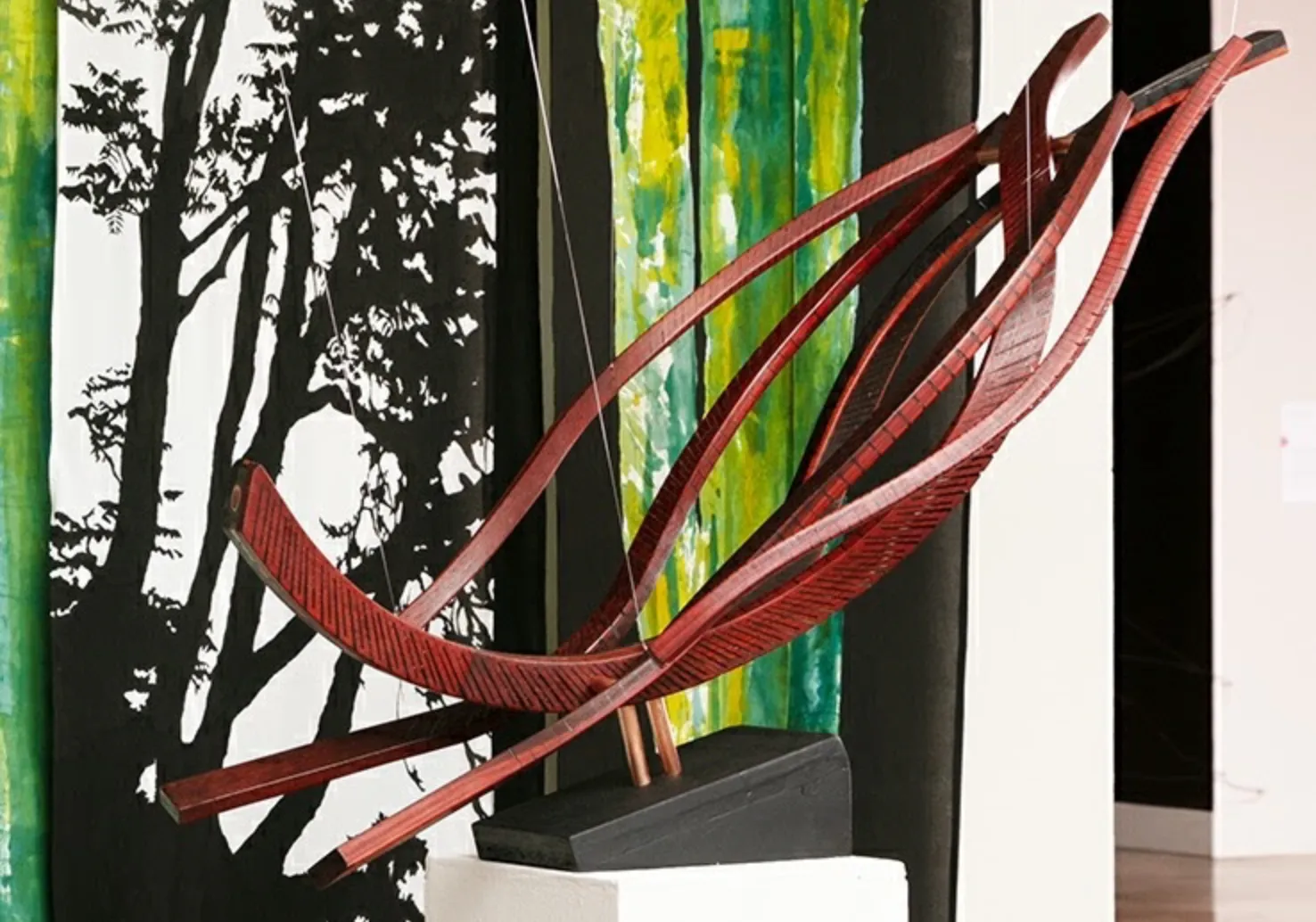 A sculptural artwork featuring curved wooden elements is displayed on a black pedestal. The sculpture is composed of several long, curved wooden strips, creating a flowing, dynamic form. The background includes abstract green and yellow vertical stripes on one side and a black and white silhouette of tree branches on the other, adding contrast to the natural wood tones of the sculpture.