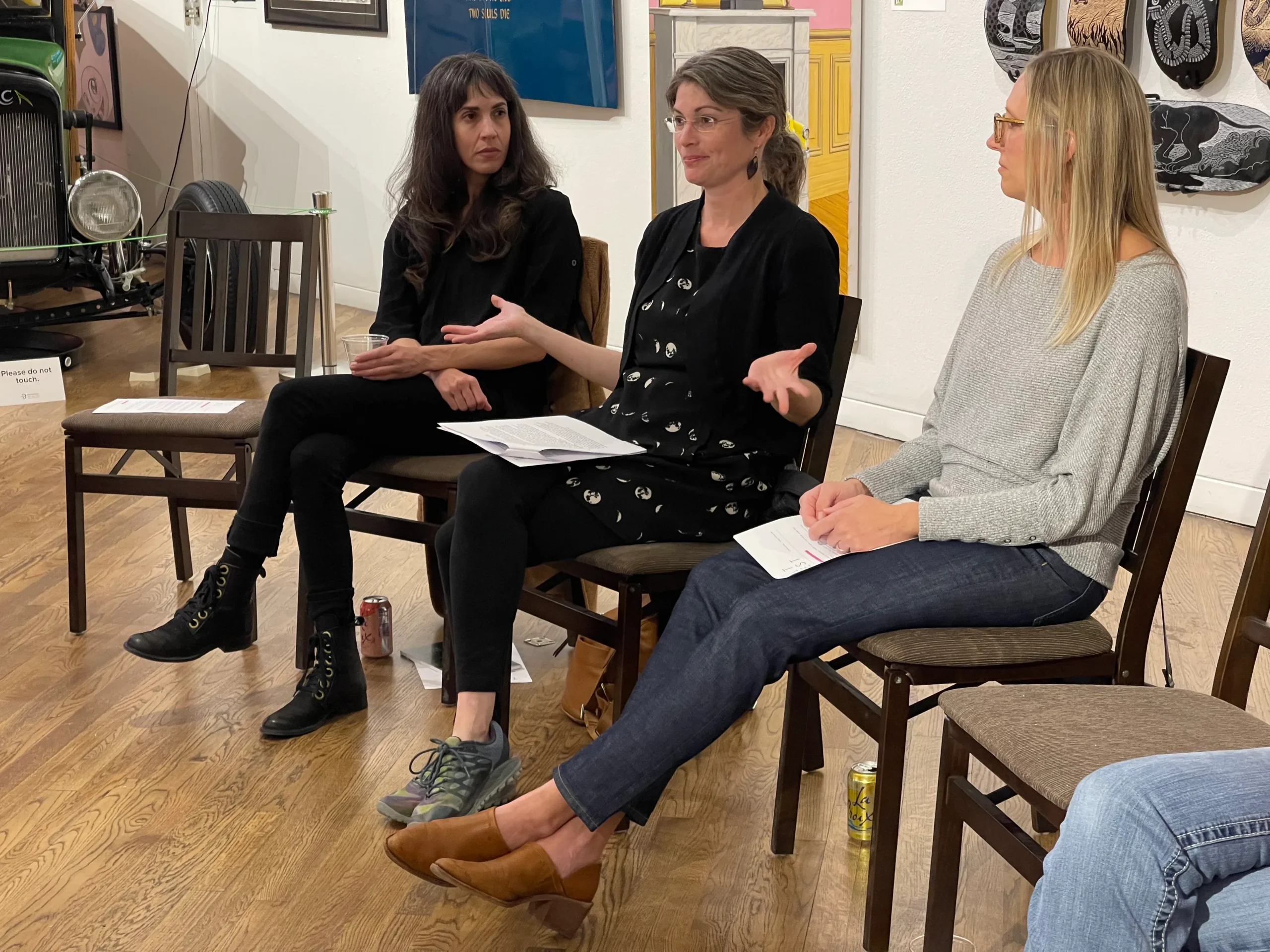 Three women sit in chairs arranged in a row, engaged in a discussion. The woman in the middle is gesturing with her hands as she speaks, while the other two listen attentively. The room features a wooden floor and artwork on the walls, including a vintage car partially visible in the background.