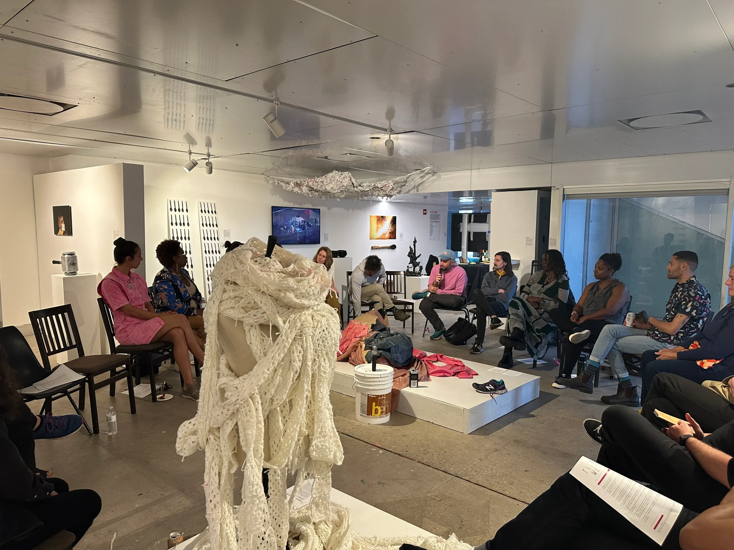 A group of people gather in chairs in a roundtable format in a white room with floor to ceiling windows. They wear bright pinks and dark colors. In the center of the table and overhead are tapestry-based sculptures. IN the front is a large white knitted sculpture that appears as an abstracted human form.