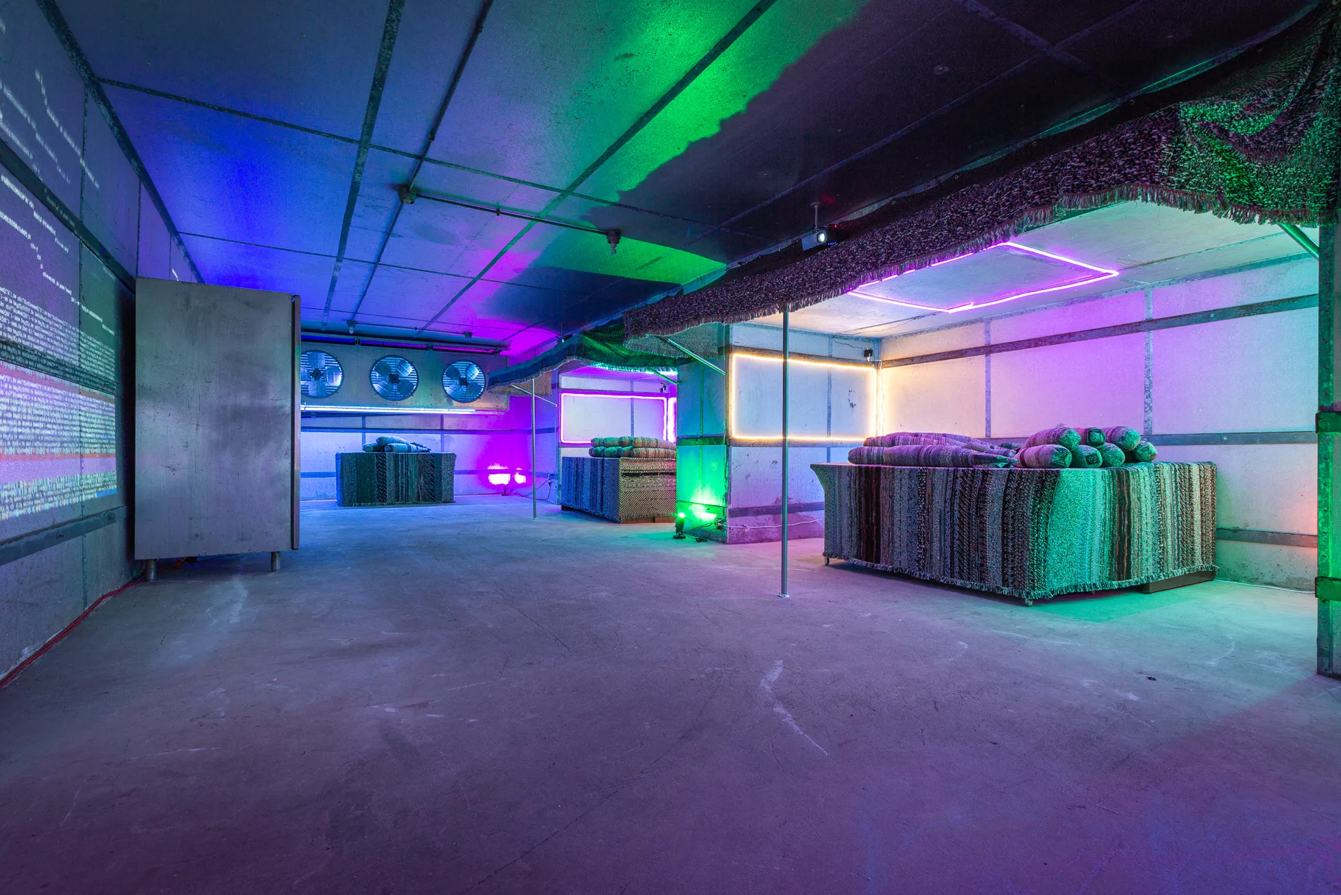 A multimedia art installation in which lighting casts bright blue, bright green and bright pink flashes on a grey-black warehouse space. There are computers, round dials on the wall, tapestries that look like bombs on a table and colorful computer-generated scripted shapes across one wall.