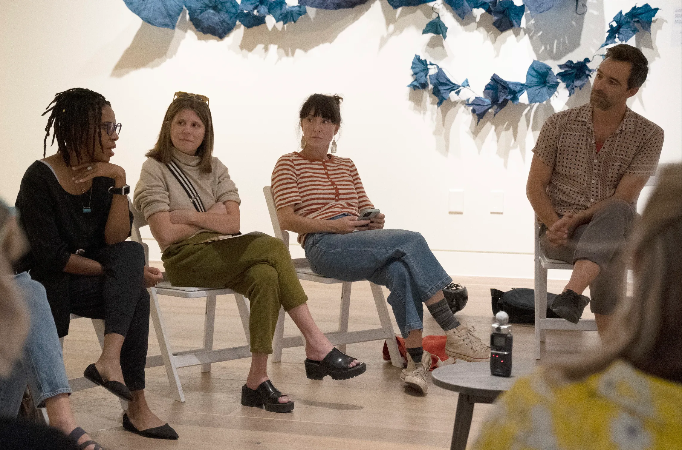 A group of people sit in a semi-circle in a gallery space, engaged in a conversation. The person speaking on the left is wearing glasses and a black outfit. The others listen attentively. The background features a wall adorned with large blue abstract fabric art.