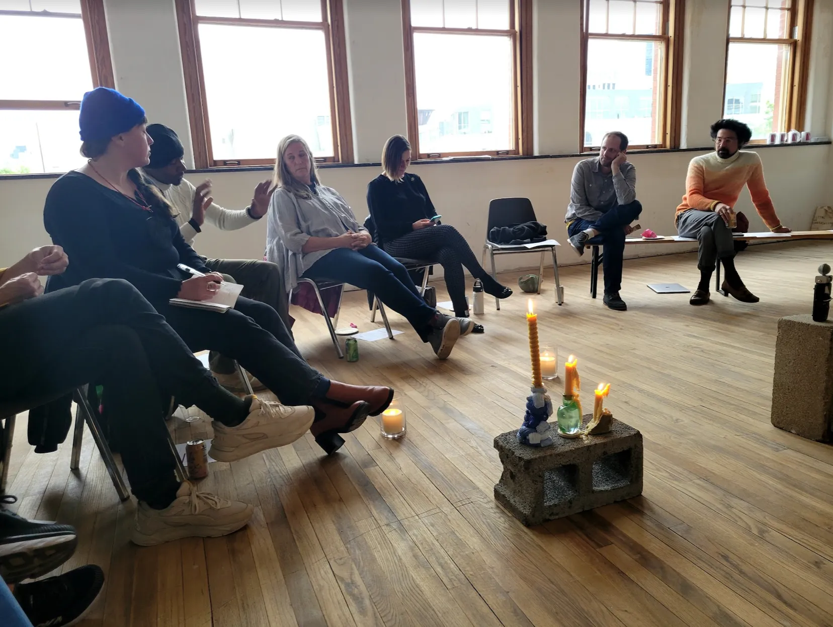 A group of people sit in a circle in a spacious room with wooden flooring and large windows. The discussion appears to be ongoing, with some participants taking notes while others listen or speak.