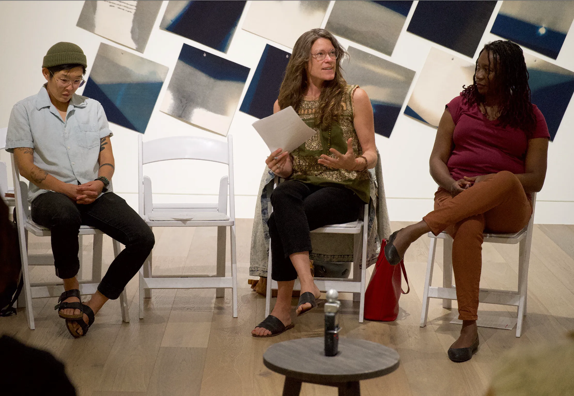 Three people sit in a gallery space, engaged in a conversation. The person in the center, holding a paper, is speaking with animated gestures, while the others listen attentively. The background features a wall adorned with abstract blue and gray square artworks.