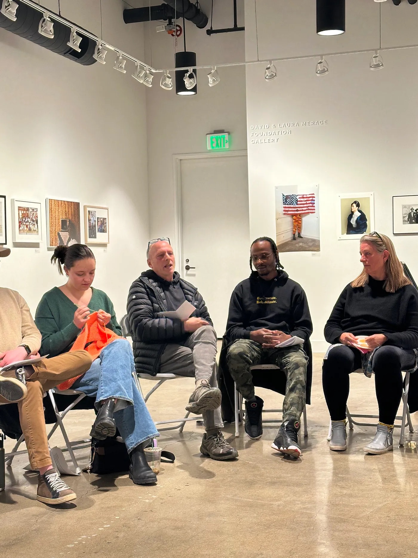 A group of people sit in a circle in a gallery space, engaged in a discussion. Some are holding knitting materials, and one person is knitting a bright orange piece. The walls are decorated with framed artwork, and the room is brightly lit with overhead track lighting.