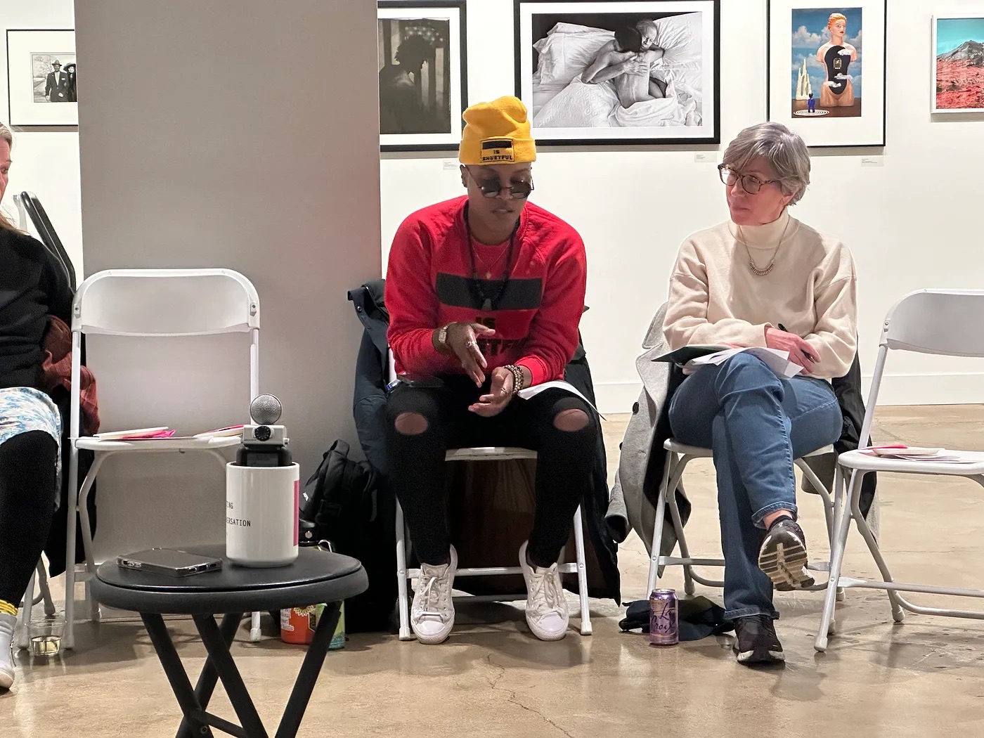 Two people sit in a gallery space, engaged in conversation. The person on the left is wearing a yellow beanie, sunglasses, a red sweater, and ripped jeans, while the person on the right is wearing glasses, a beige sweater, and jeans. The background features framed photographs on the wall.