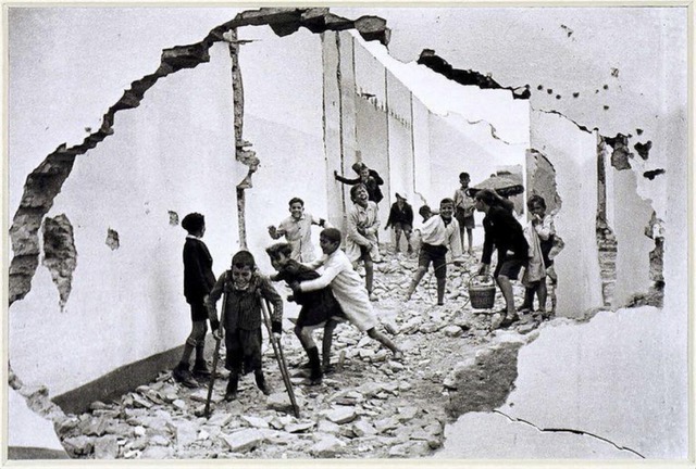 A black and white photograph of eleven children playing in the rubble of a destroyed building. The photo is taken through the view of a hole in the wall. The child in the forefront balances on crutches