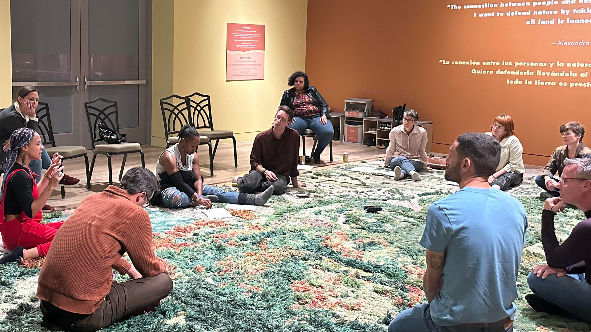 A group sits in a circle, some on black chairs, most on a colorful rug-like artwork in green, yellow, and burnt orange extending up one wall. Other walls are yellow and orange, with artwork and painted text.