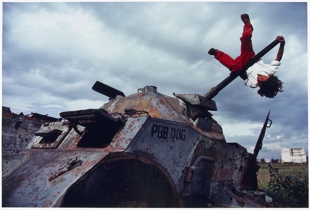 A color photograph in which a child with long, curly black hair wearing red pants and a white shirt swings upside down on the turret of a rusted war tank. A cloud-filled sky and the rubble of buildings hover in the background