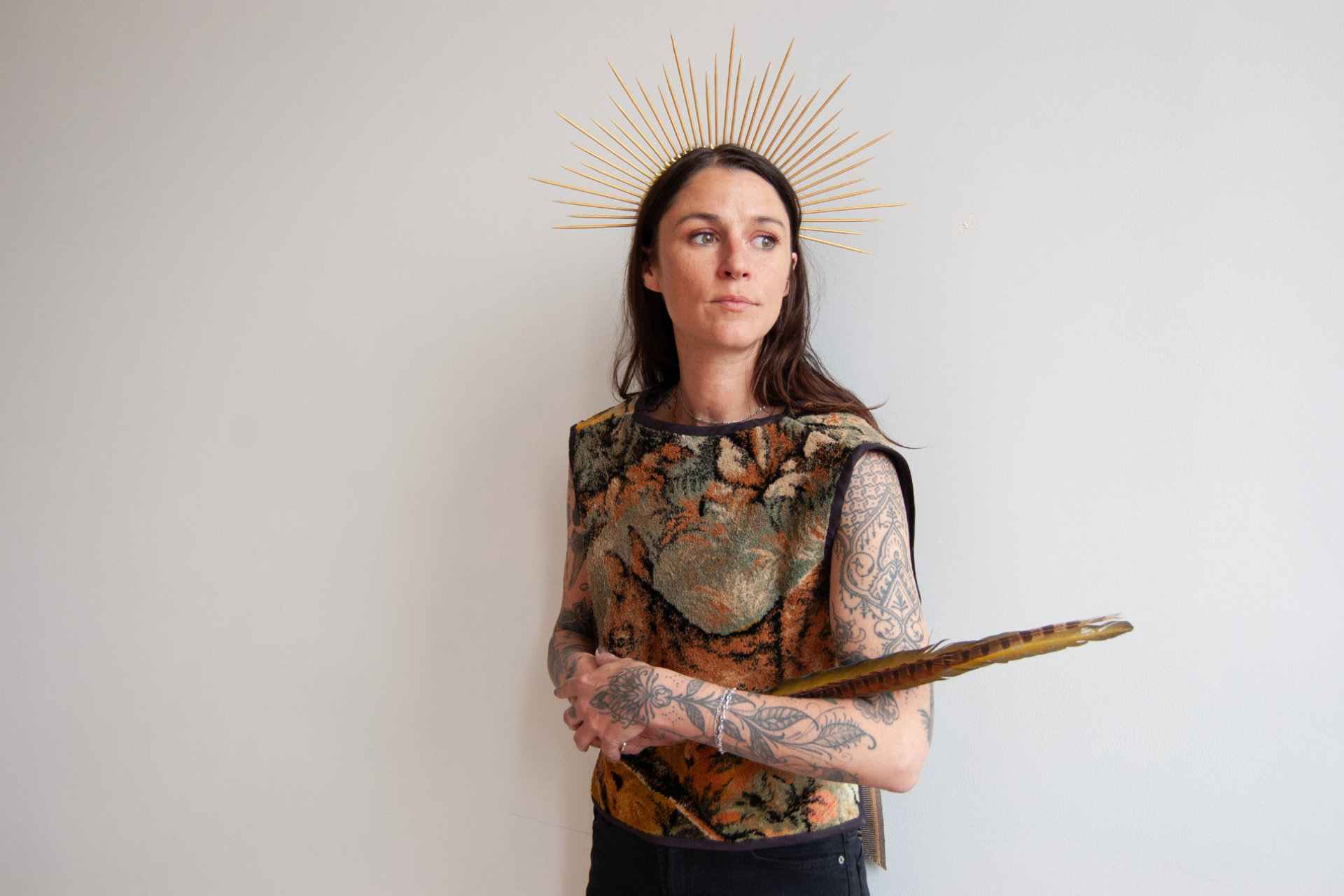 Woman holding a large feather, wearing a tapestry vest and a spiked wooden crown.