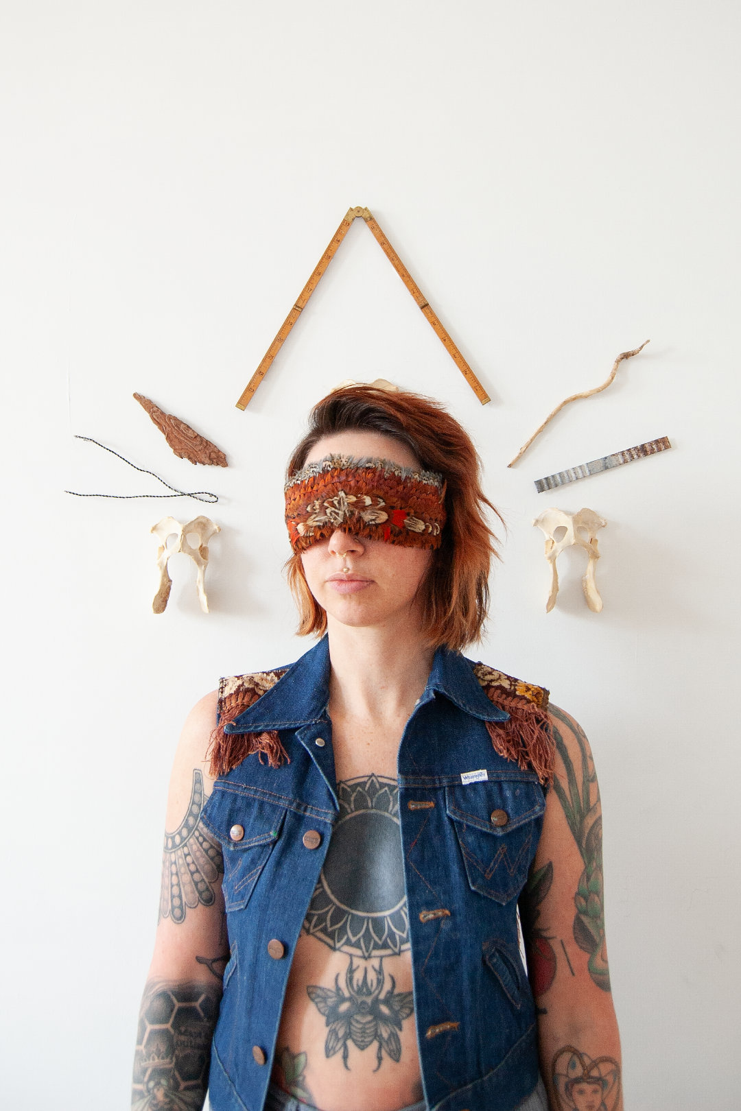 Woman with a feather blindfold, tattoos visible, standing before mounted objects like bones and sticks.