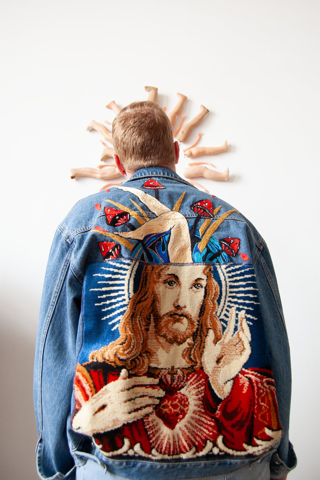 Man’s back facing the viewer, framed by a circle of doll legs, wearing a denim jacket with tapestry art.