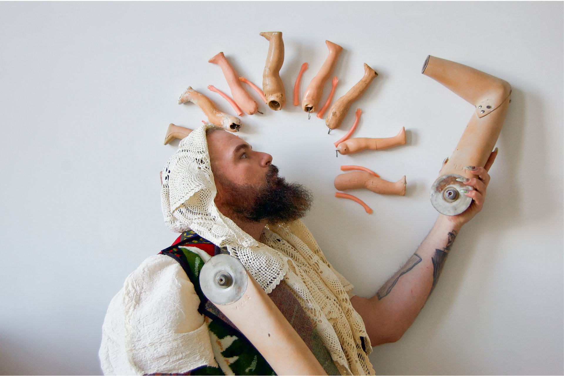 Profile of bearded figure with a lace doily on head, holding a mannequin arm, doll limbs mounted on the wall.