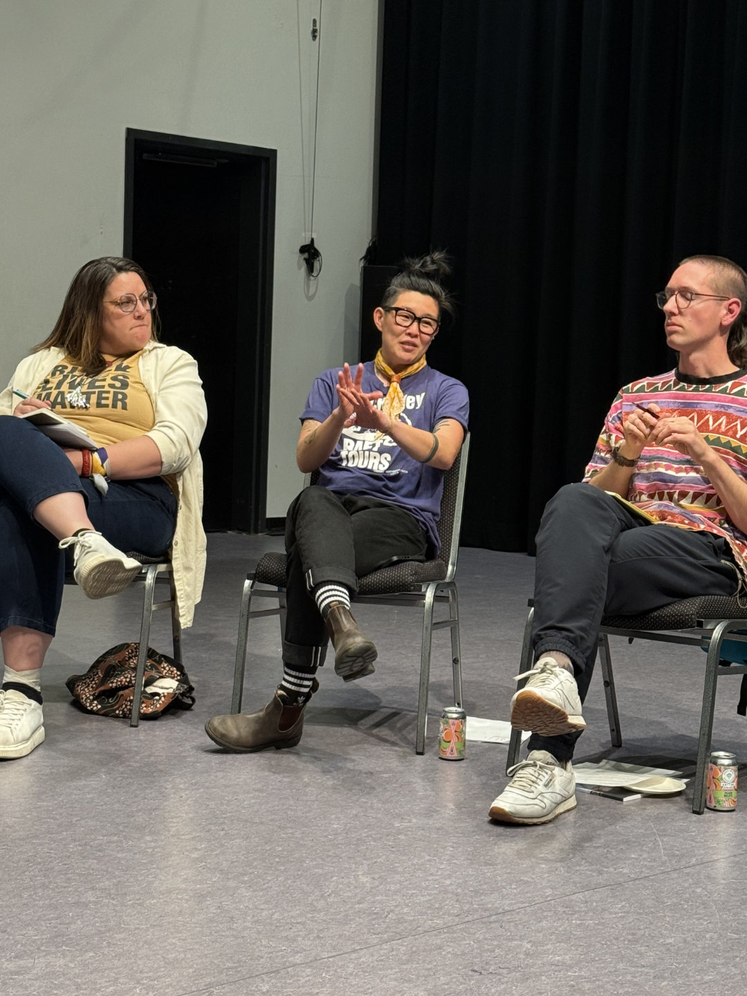 Three people seated in a row indoors; the person in the center speaks while gesturing with their hands